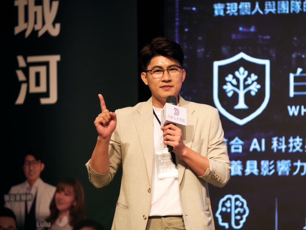 Man in a beige suit speaks at a conference, holding a microphone and raising one finger.