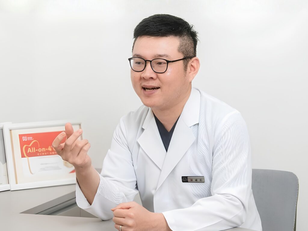 Man in a white lab coat and glasses speaks, using hand gestures in a clinical setting.