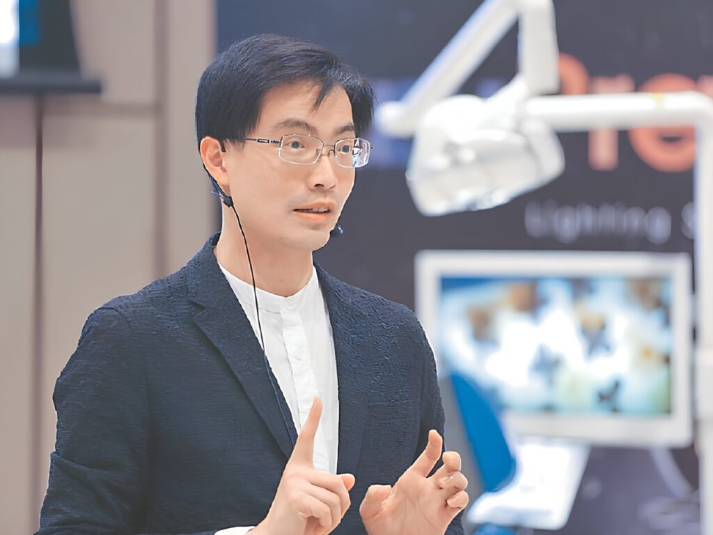 Man with glasses and a headset presenting, gesturing with both hands in front of a blurred display behind him.