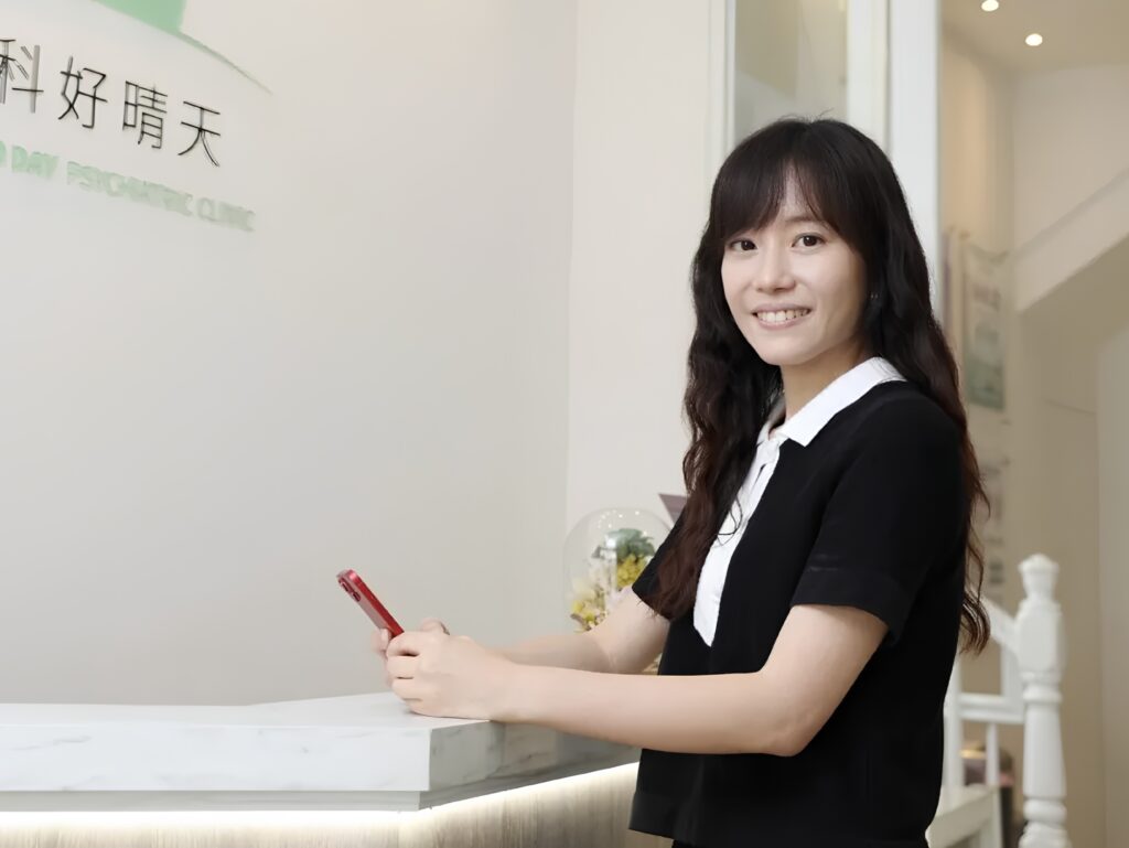 Woman in a black uniform standing at a marble reception desk, smiling at the camera while holding a red pen.]',']}{